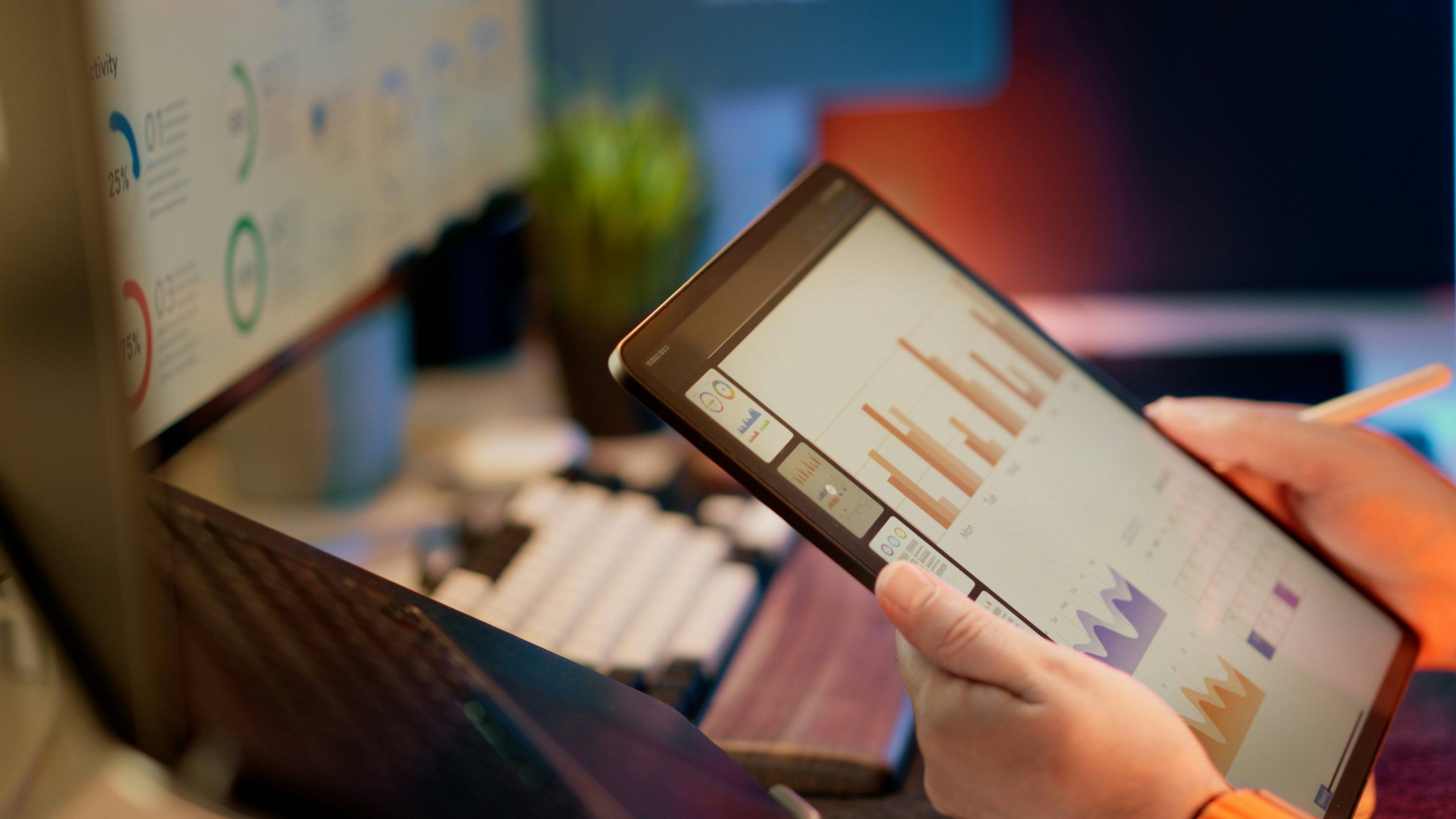 Person reviewing charts on a tablet beside a monitor.